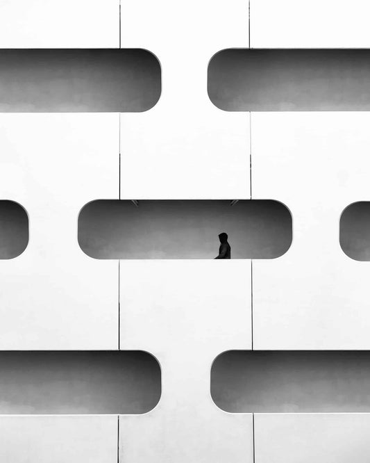 The artpiece 'Boxed' by Nina Papiorek shows a beautiful facade of a building at the university of Paris, with one lonely hooded person walking through one of the 'frames' created by the cutouts of the facade. The artwork is completely in black and white.