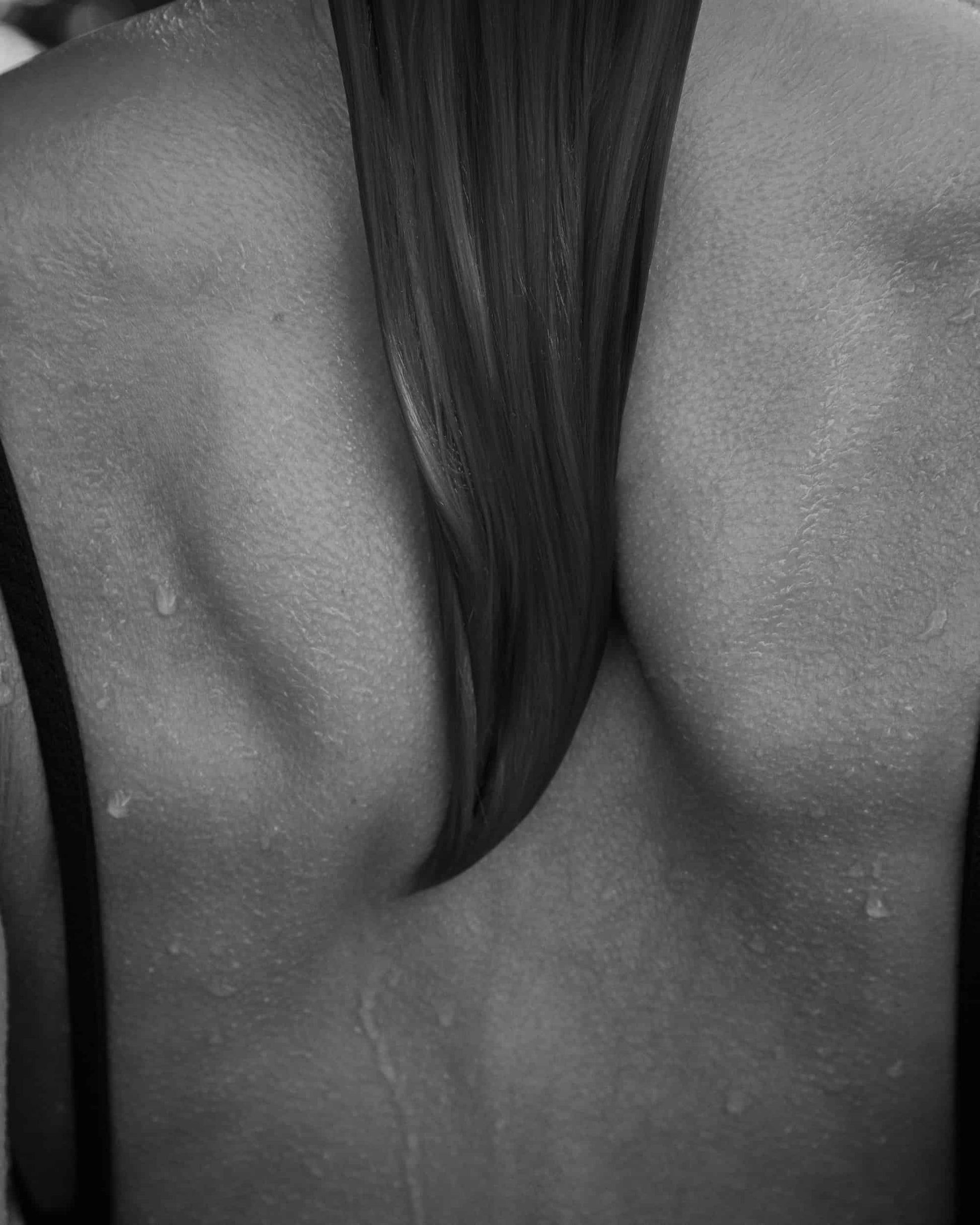 The artpiece 'Be' by Andreas Ortner shows a bare back with waterdrops on it and long hair hanging inbetween the shoulderblades, in black and white.