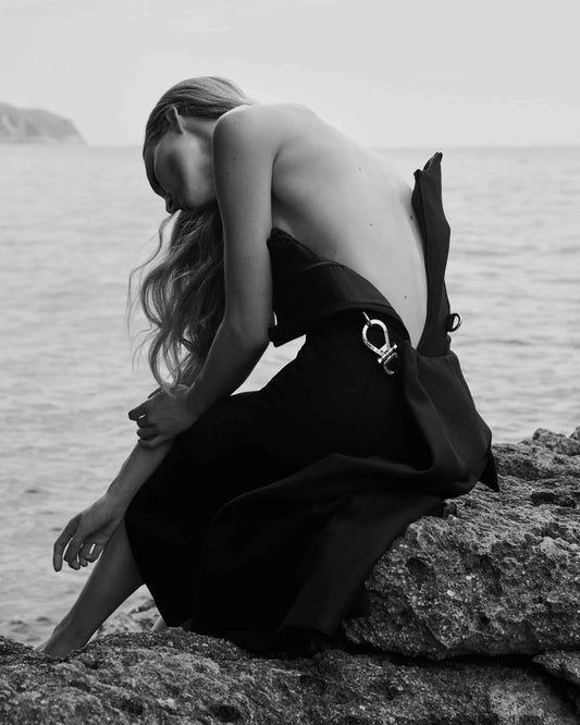 The artpiece 'Deep' by Andreas Ortner shows a woman sitting on rocks next to the sea with a backless dress and her hair draped down, she is slightly bending her back downwards and laying her hand on her own arm.