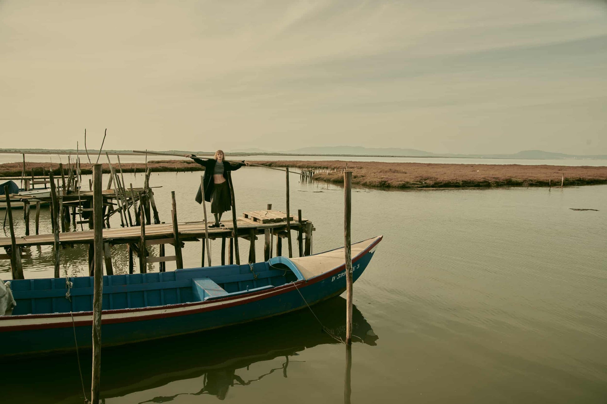 The artpiece 'Embrace' by Andreas Ortner shows a few wooden piers in a lake with a blue fishing boat in the front. A woman is standing on a pier with her arms widespread.