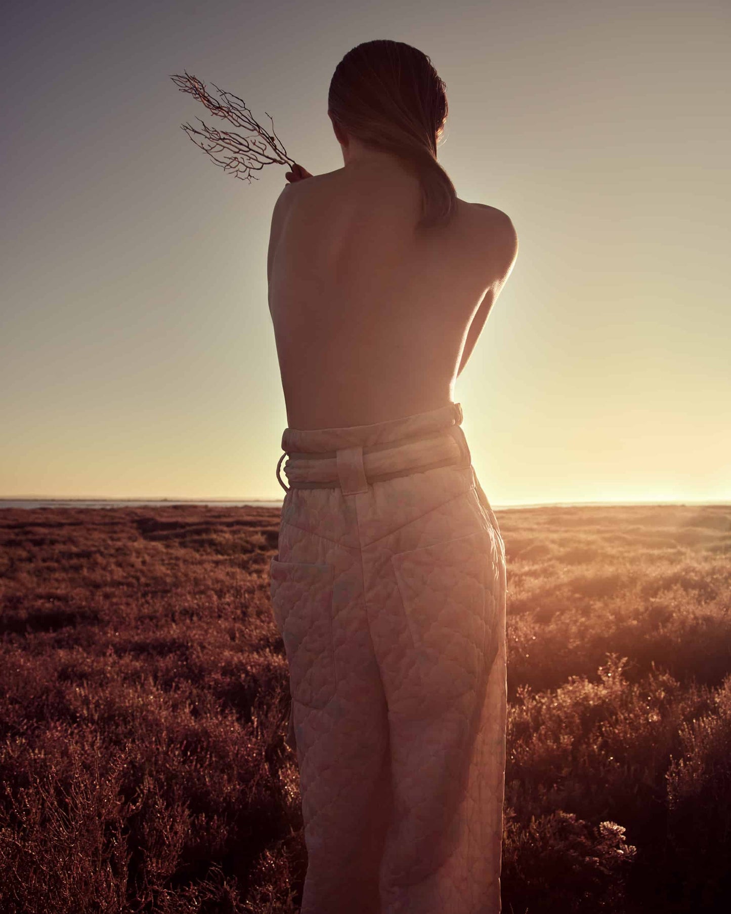The artpiece 'Hold' by Andreas Ortner shows a woman in a long grass field with the sunset at the horizon, who is wearing high-waisted pants and no top. She is embracing herself and holding a twig in one hand.