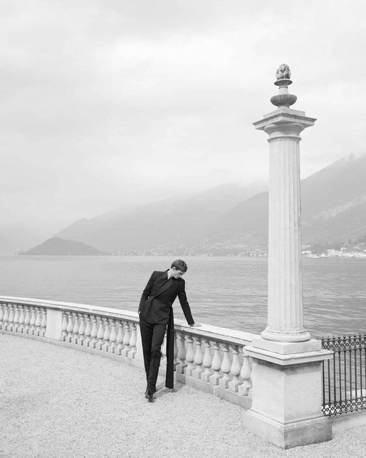 The artpiece 'Present' by Andreas Ortner shows a man in a suit leaning against a stone railing in front of a lake, next to a greek style pillar.