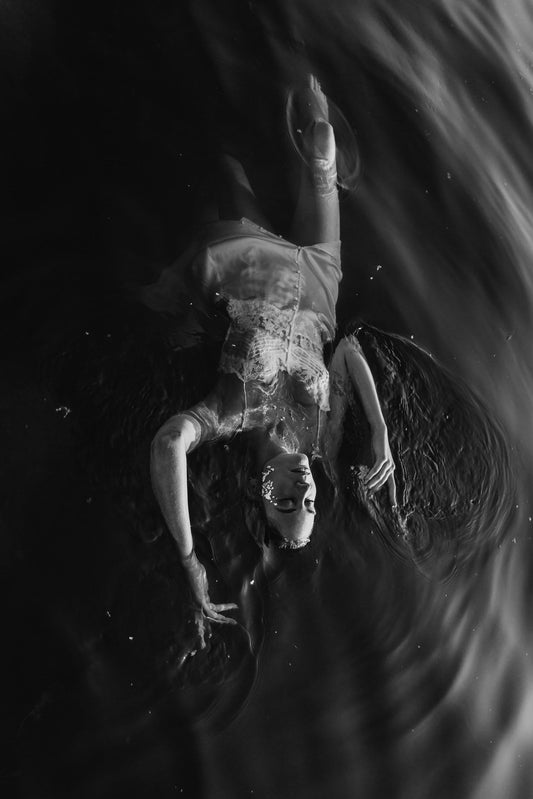 Human form floating in dark waters, reflecting life's fluidity, in high contrast black and white photography by Celin May.