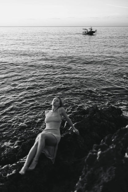 Woman reclining on rocky shore with a distant boat, captured in a thoughtful black and white photograph by Celin May.