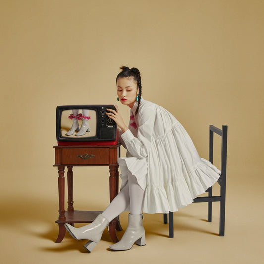 The artpiece 'Desired' by Hui Long shows a tv with an image of a pair of ankle boots wrapped with red flowers, and a woman next to the tv, who is wearing the ankle boots, but without the flowers.