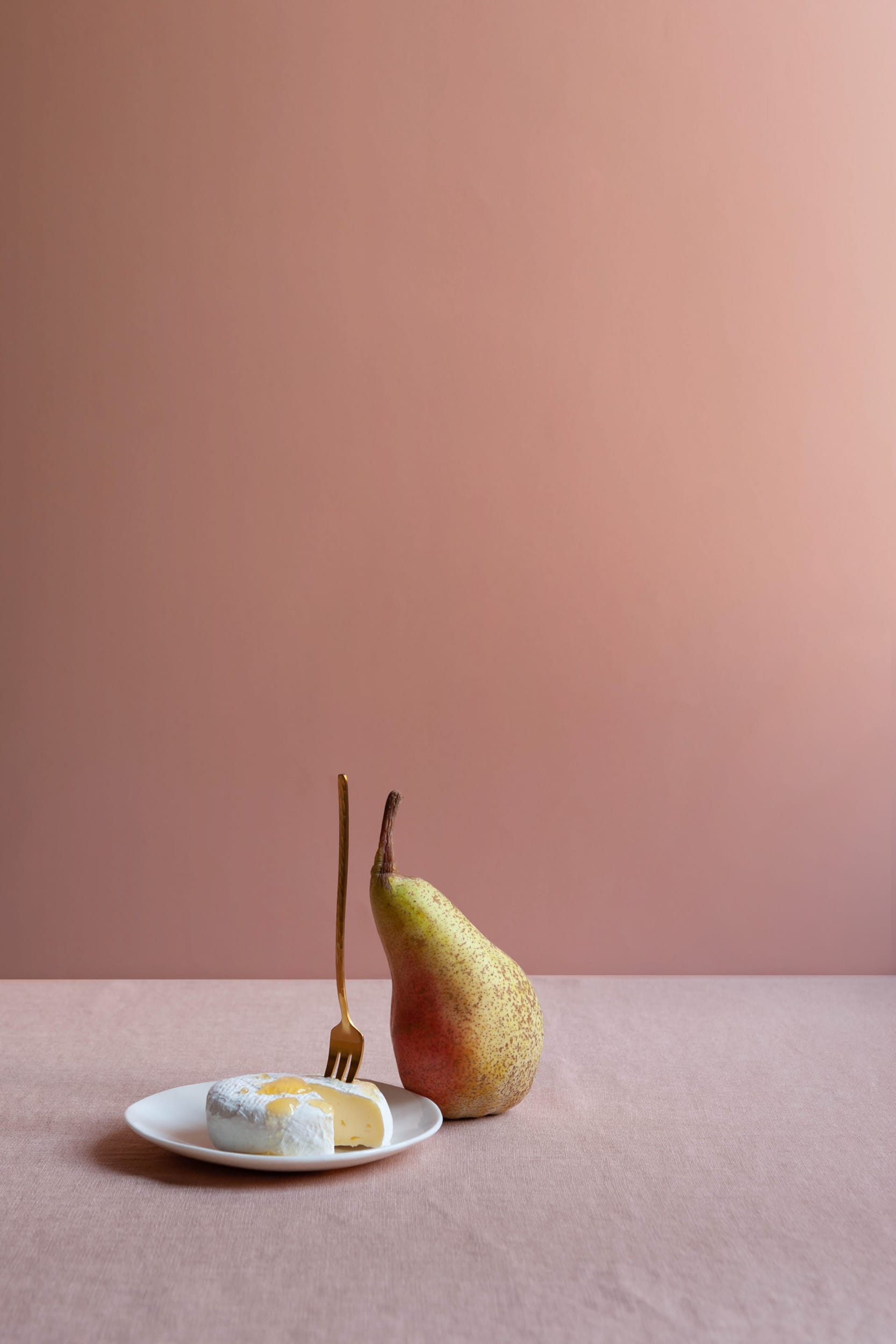 The artpiece 'Poise' by Erica Ferraroni shows a still life photo, with all objects in perfect equilibrium. A golden fork, a pear and a honey drizzled cheese laid out against a pink background.