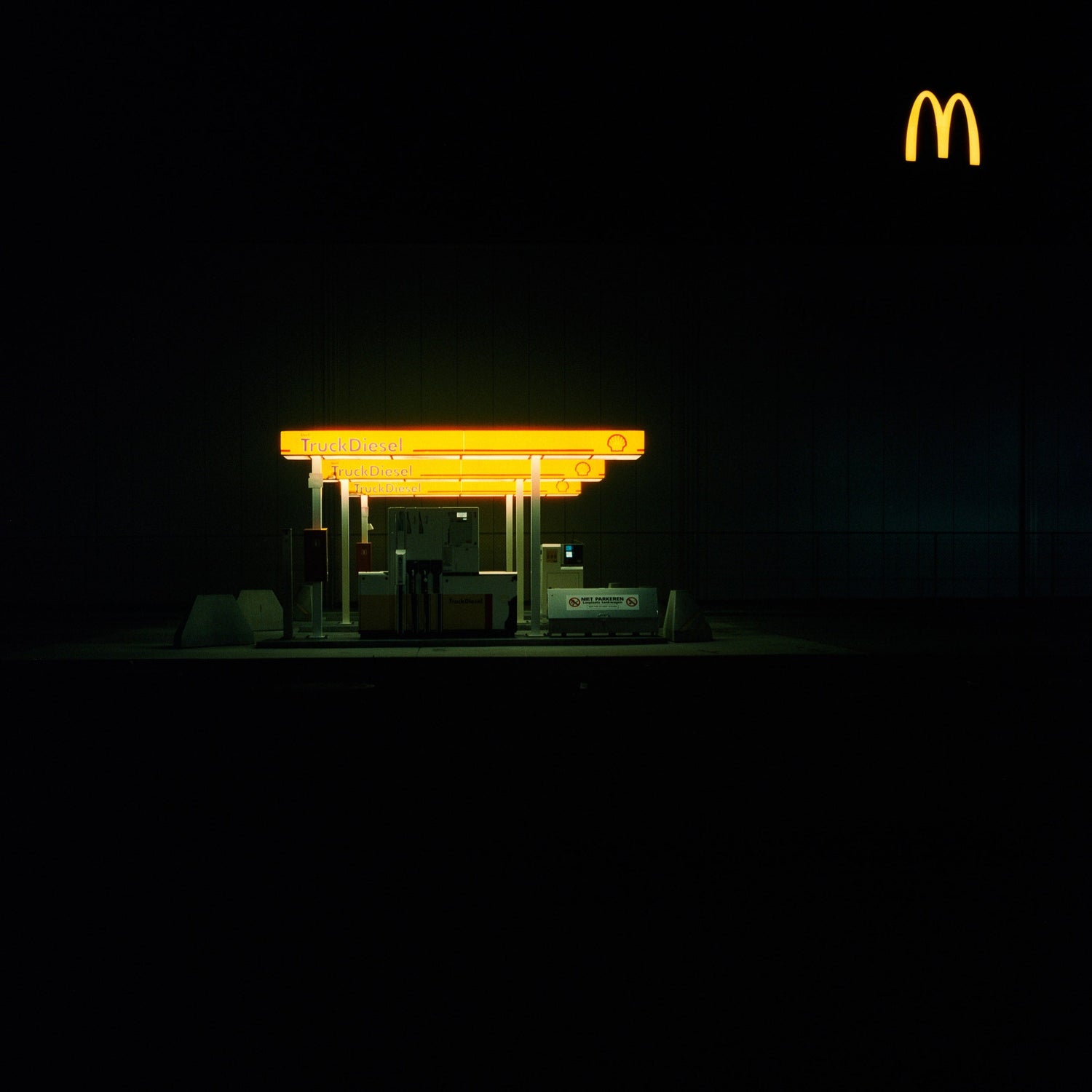 The artwork 'Exterior #9' by Jildo Tim Hof displaying a truck gas station lit up by bright yellow lights against a dark sky