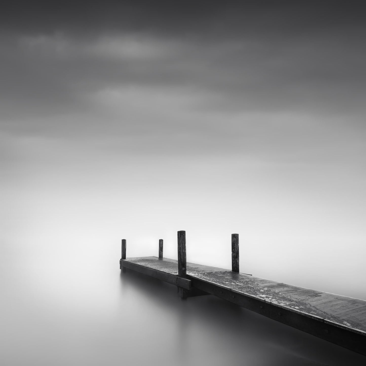 The artpiece 'Shadows of Silence' by Marco Maljaars shows a wooden catwalk, on top of a silent mysterious lake. Shot in black and white.