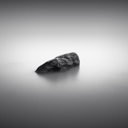 The artpiece 'Simplicity II' by Marco Maljaars shows the top of a rock laying in a quiet and silent lake. Shot in black and white.