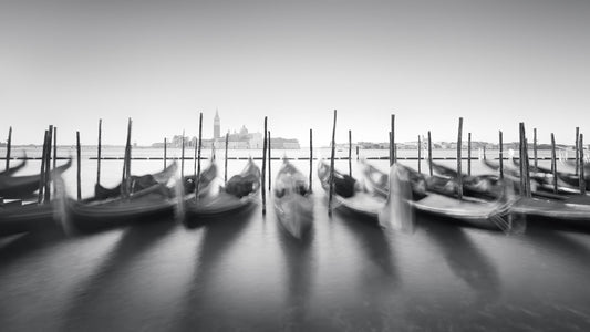 The artpiece 'Dancing Gondola' by Marco Maljaars shows a long exposure black and white image of moored gondola's in Venice, with the city visible in the background.