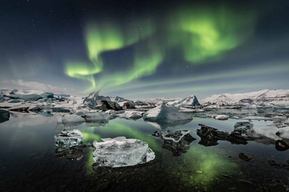 The artpiece 'Jokulsarlon' by Markus van Hauten shows a rocky snowy lake, with a bright green aurora in the sky and reflecting in the water.