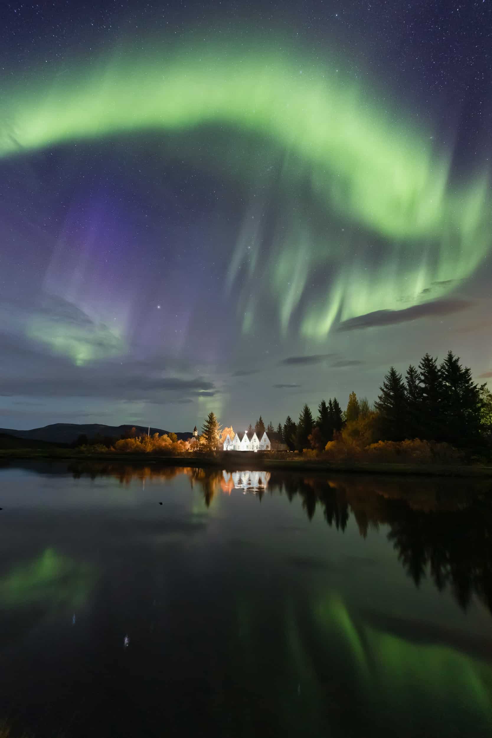 The artpiece 'Vatn' by Markus van Hauten shows a bright green and purple aurora over a lake and a small settlement on the lake edge.