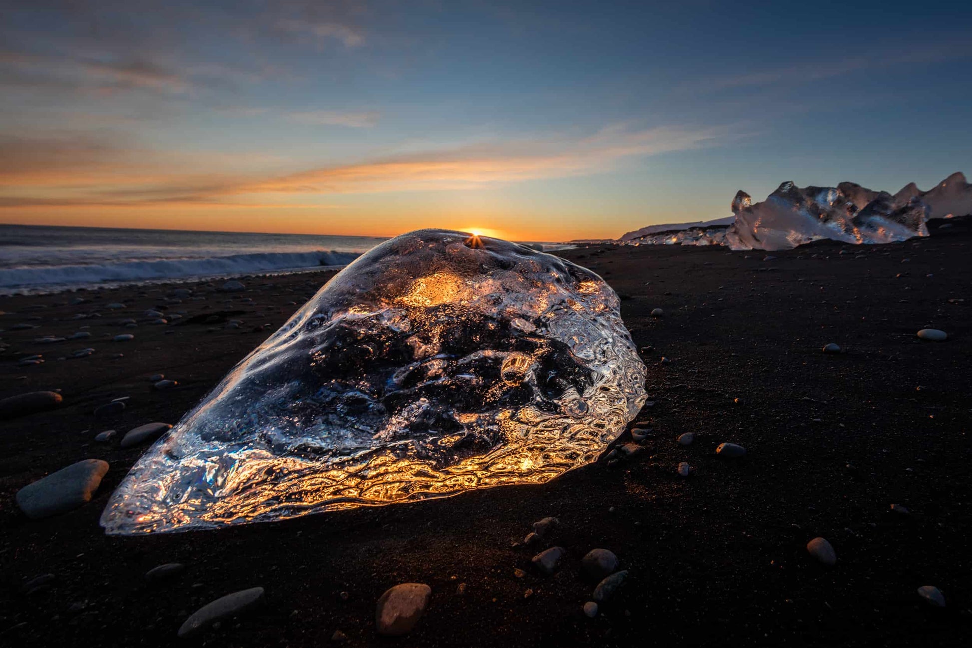 The artpiece 'Ice Diamond' by Markus van Hauten showing an ice rock in the shape of a diamond on a beach at dawn, scattering the light through the ice.