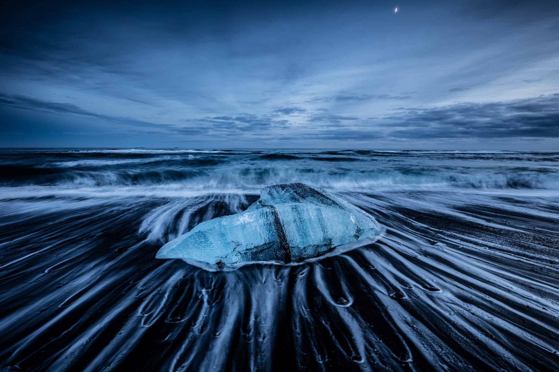 The artpiece 'Satin Blue' by Markus van Hauten which shows an ice rock, sitting in the surf at night, with waves retreating to the ocean around it.