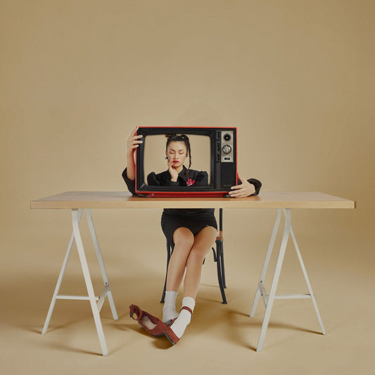 The artpiece 'Mask' by Hui Long shows a person sitting at a desk, their upper body hidden behind the desk and a tv which is standing on the desk. The TV shows an upper body of a woman, who may or may not be the person sitting at the desk.