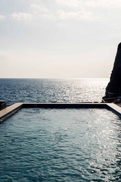 The artpiece 'Piscine Avec Vue' by Mathieu Puga shows a pool on the edge of the ocean, with a rocky hill coming up on the side.