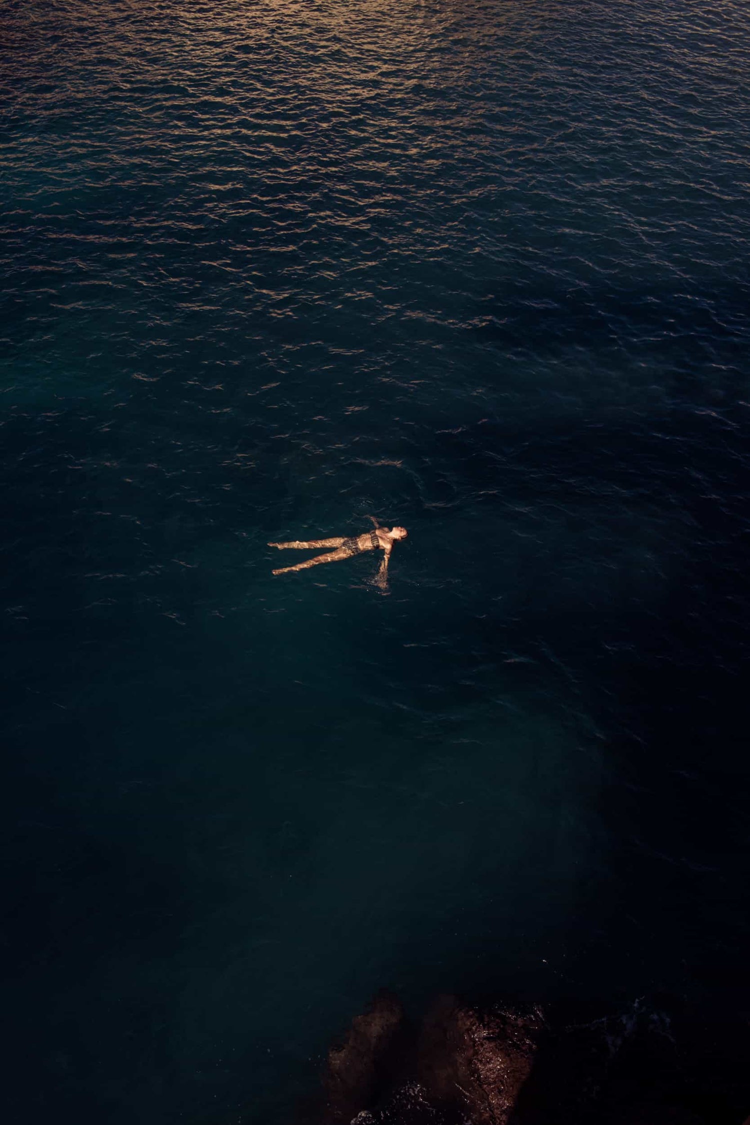 The artpiece 'Home Again' by Mathieu Puga is a black and white photograph which shows a person floating in a dark blue ocean, while the sunlight is reflecting on the waves in the background.