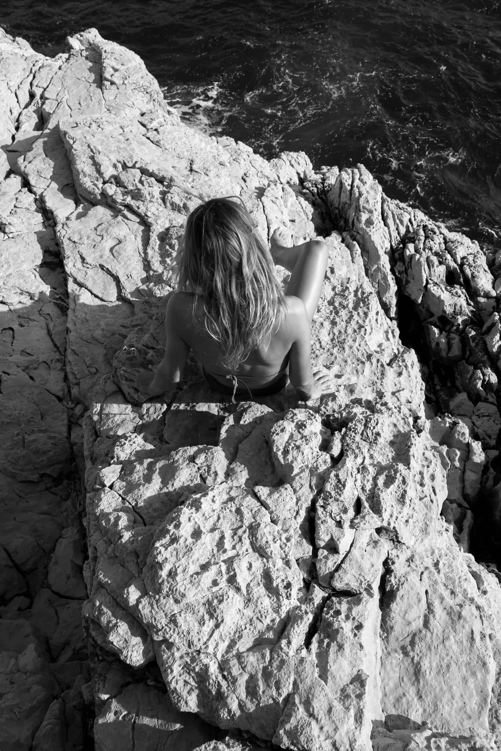 The artpiece 'Plongée' by Mathieu Puga is a black and white fine art piece, an oceanfront image, with a woman, imaged from above sitting on the rocks at the waters edge.