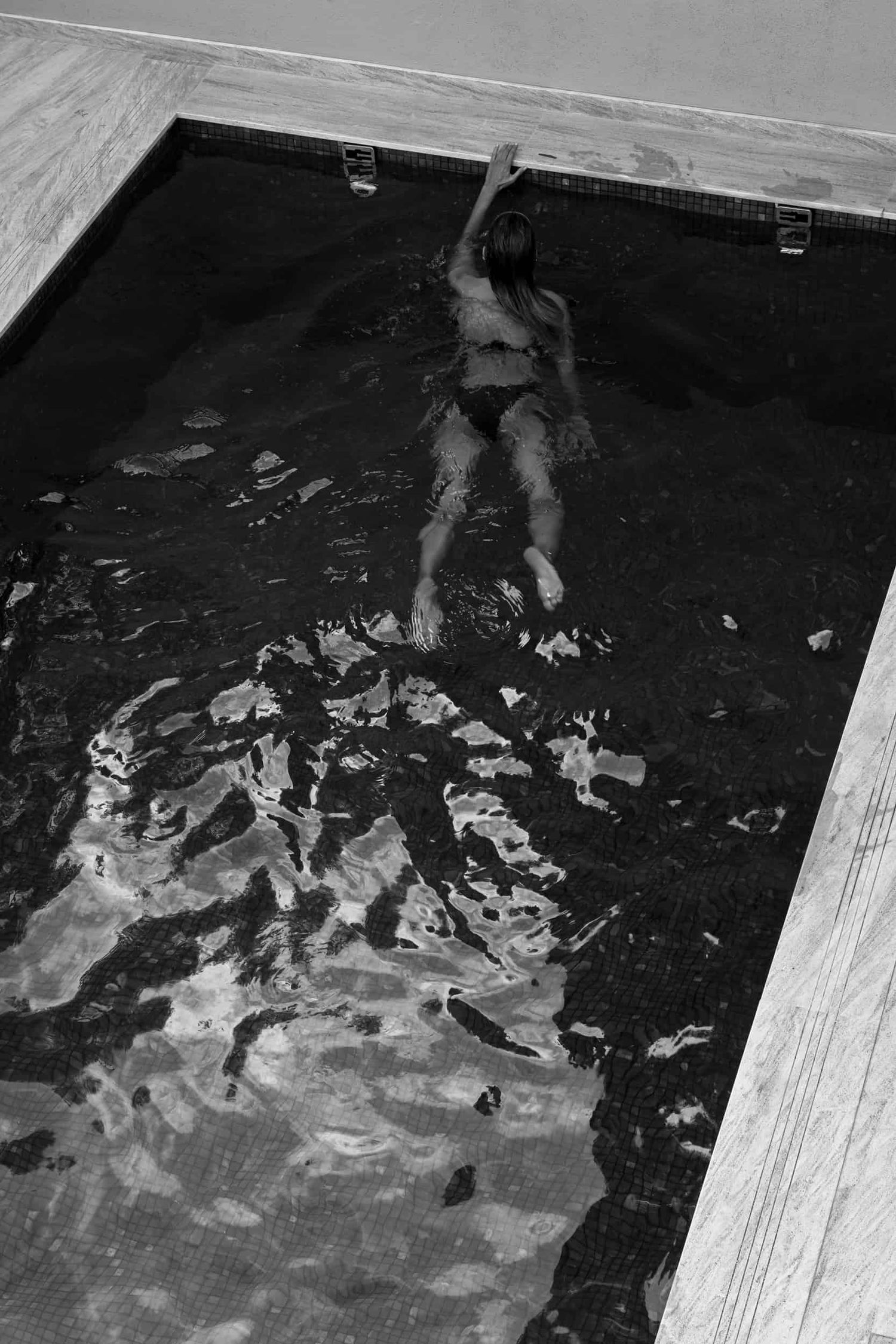 The artpiece 'She Was Here' by Mathieu Puga is a black and white fine art piece and pictures a woman swimming in a pool, reaching for the edge, seen from above.