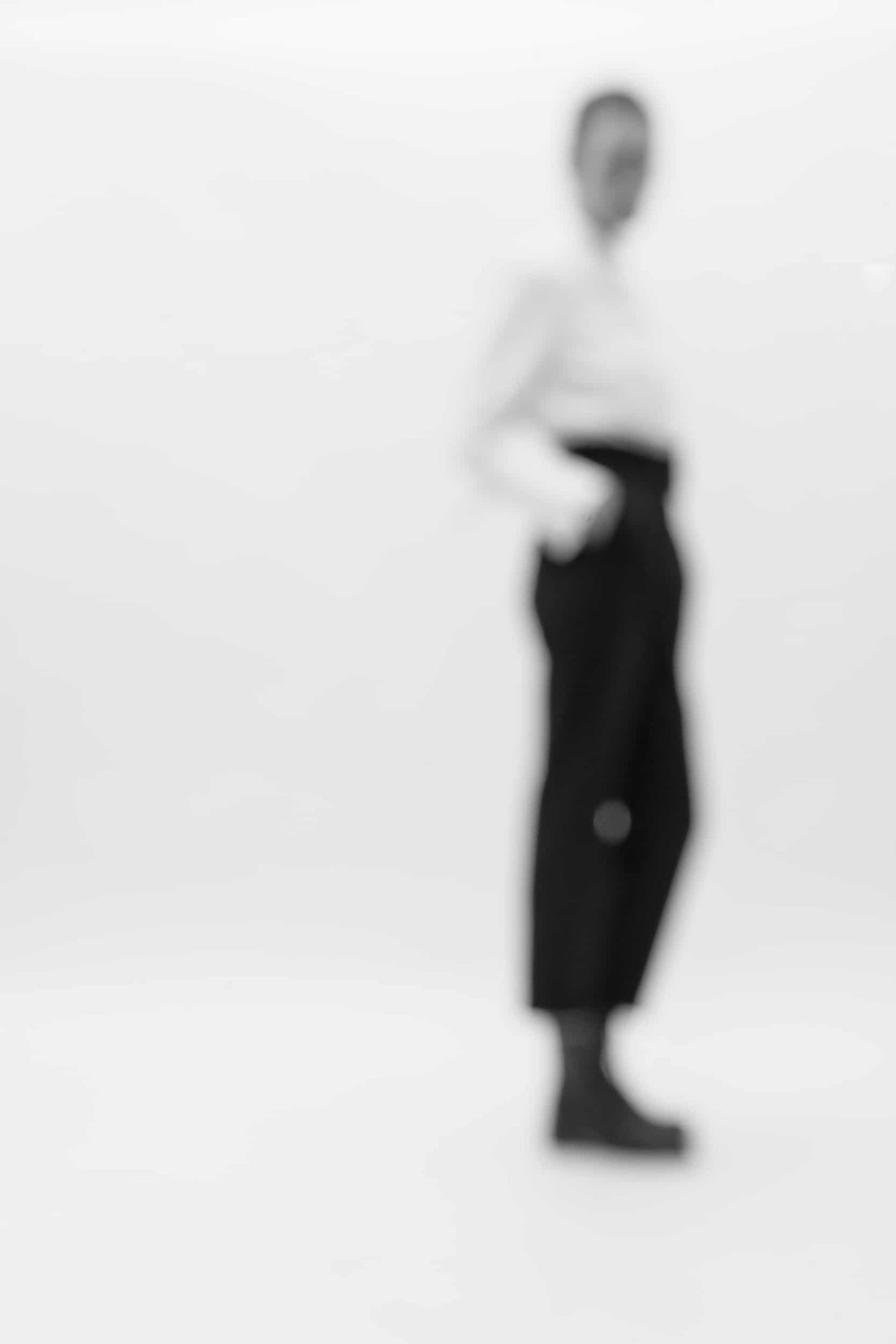 The artpiece 'Silhouette' by Mathieu Puga is black and white fine-art piece, which shows a woman in black pants and a white blouse, standing with her hand in her pocket. The image is blurred, causing parts of the image to seemingly disappear into the background.