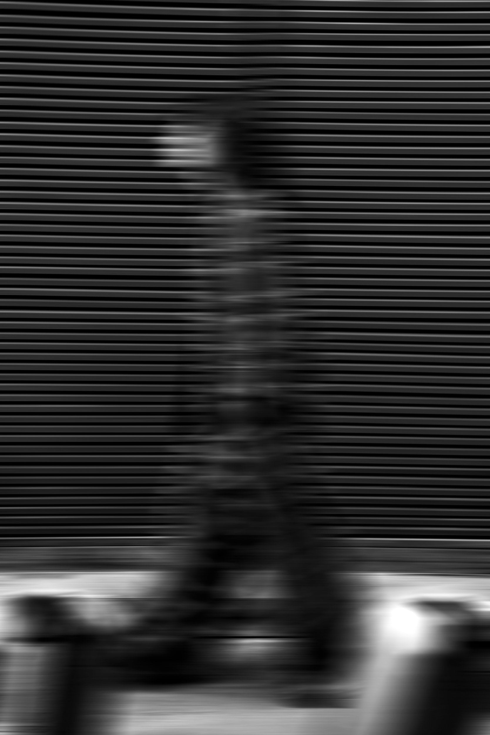 The artpiece 'Passenger' by Mathieu Puga shows a faded person walking in front of a patterned wall, with horizontal stripes visible through the blurry person. Creating an image seemingly full of motion.