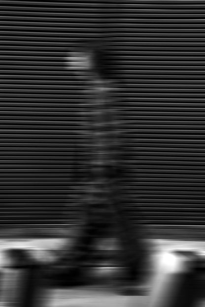 The artpiece 'Passenger' by Mathieu Puga shows a faded person walking in front of a patterned wall, with horizontal stripes visible through the blurry person. Creating an image seemingly full of motion.