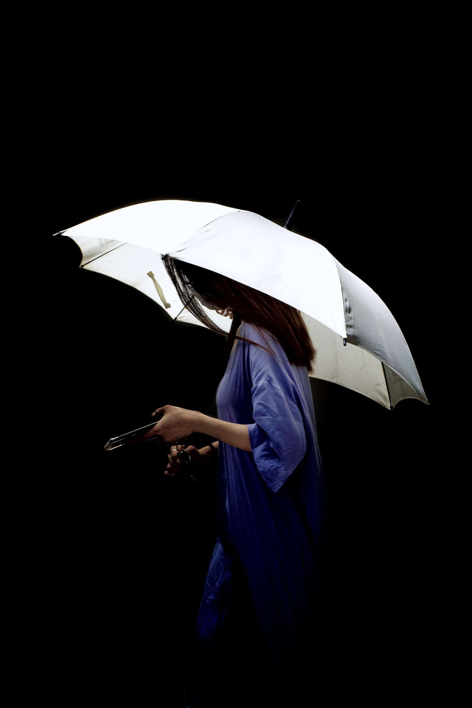 The artpiece 'Protection' by Michael Cai shows a woman in blue dress walking with a white umbrella against a completely black background.