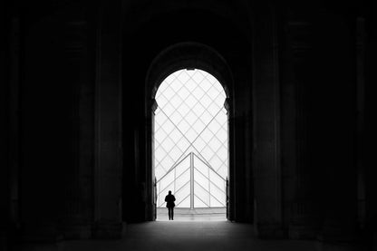 The artpiece 'Louvre' by Nina Papiorek shows a lone person standing in a doorway of the Louvre, while looking out at the iconic Louvre glass pyramids. The artwork is completely in black and white.
