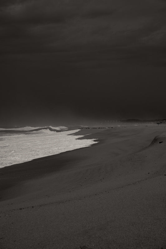 The artpiece 'Before the storm' by Rebekka Eliza shows a black and white image of a beach, with foam covered waves rolling in from the ocean and a dark and cloudy sky visible in the background.