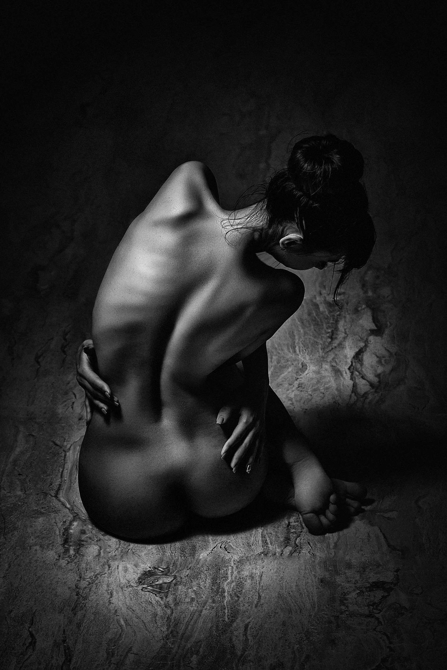Woman seated on a wood-patterned floor, her back illuminated, creating a play of lines and curves, captured in black and white by Ruslan Bolgov.