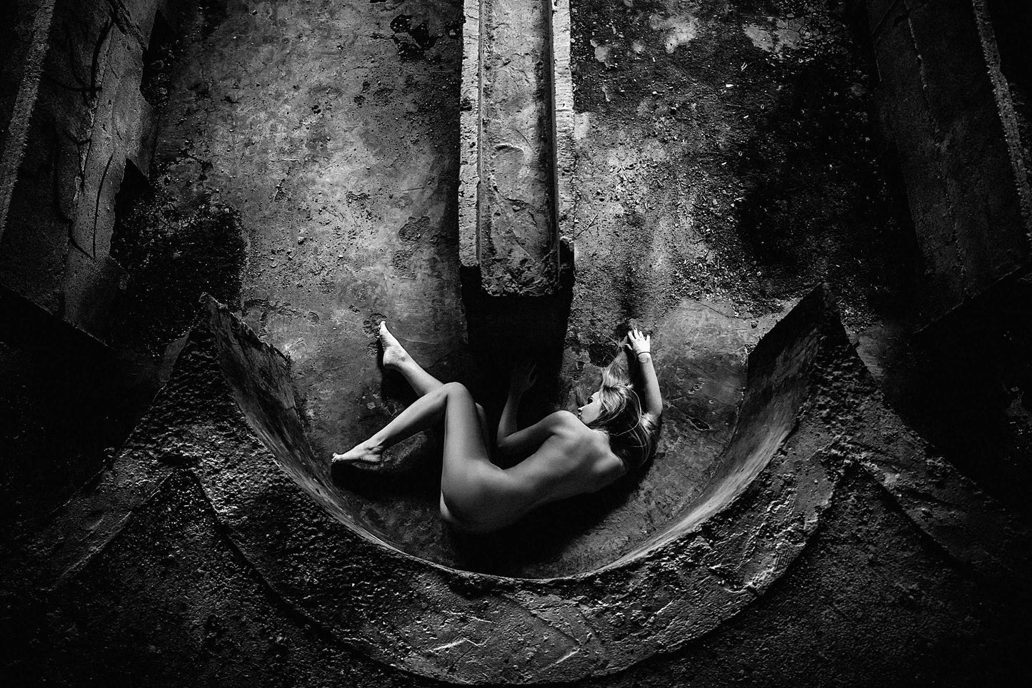 Fine artwork of a nude woman laying on her side within a curved concrete structure, captured from above, showcasing the harmony of feminine form with industrial design by Ruslan Bolgov.