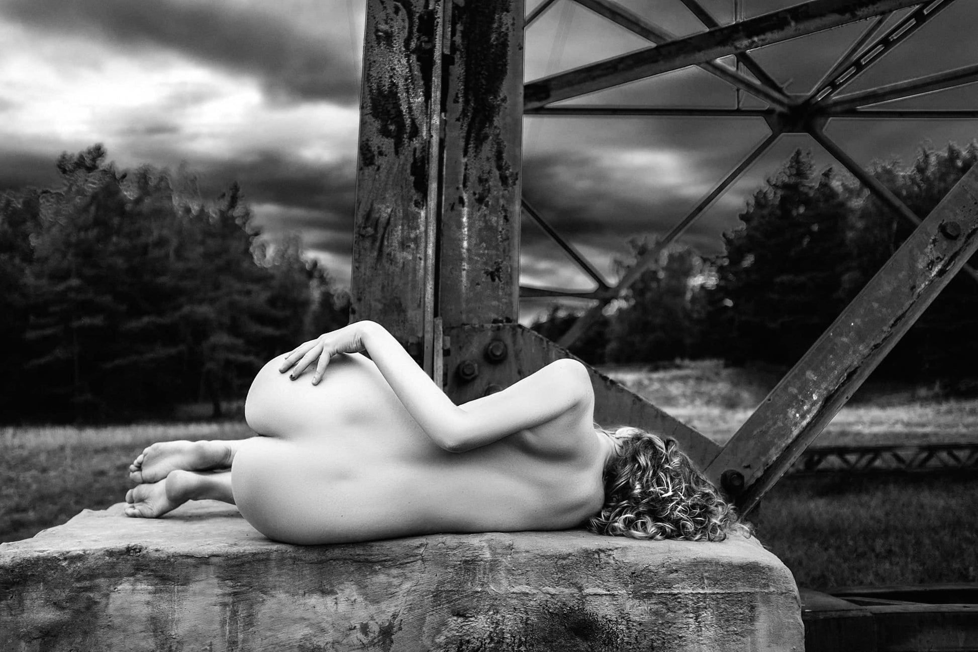 Fine artwork of a nude woman curled up on a concrete base of an electric transmission tower, with the tower's steel legs looming behind, captured by Ruslan Bolgov.