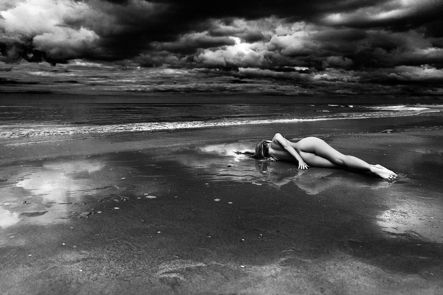 Fine artwork showing a woman laying on the beach edge, with a retreating wave glimmering around, contrasting the quiet and dark surroundings, captured by Ruslan Bolgov, available at Tones Gallery.