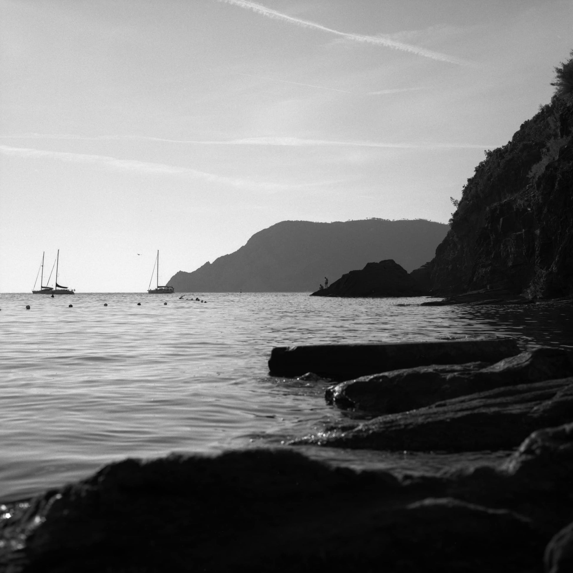 The fine-art piece 'Mar Ligure Incolore' by Tom Kluyver a rocky coastline, with two people standing on the rocks in the distance, photographed in black and white.