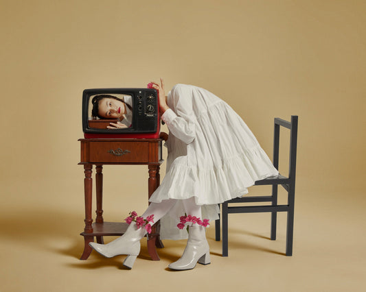 The artpiece 'Watch' by Hui Long from the collection Real and Unreal shows a woman sitting at a table while resting her head on the table behind a tv. There is a TV sitting on that table displaying the image of a woman resting her head on the same table.